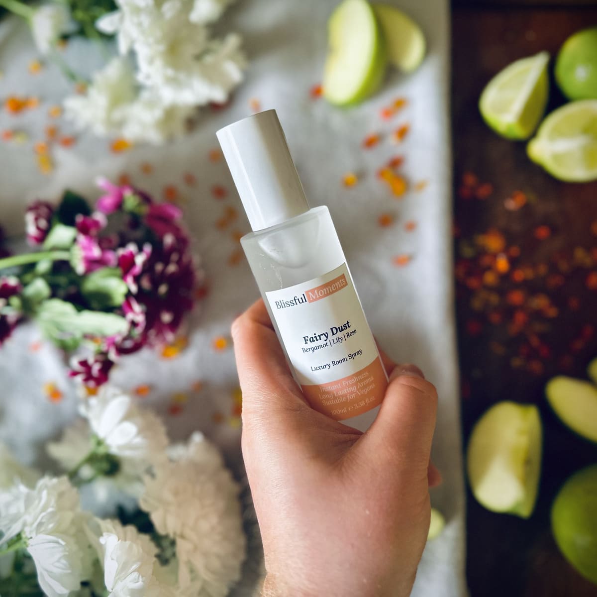 Hand holding Fairy Dust scented luxury frosted glass room spray surrounded by fresh flowers, amber and green apples sitting on a cosy blanket