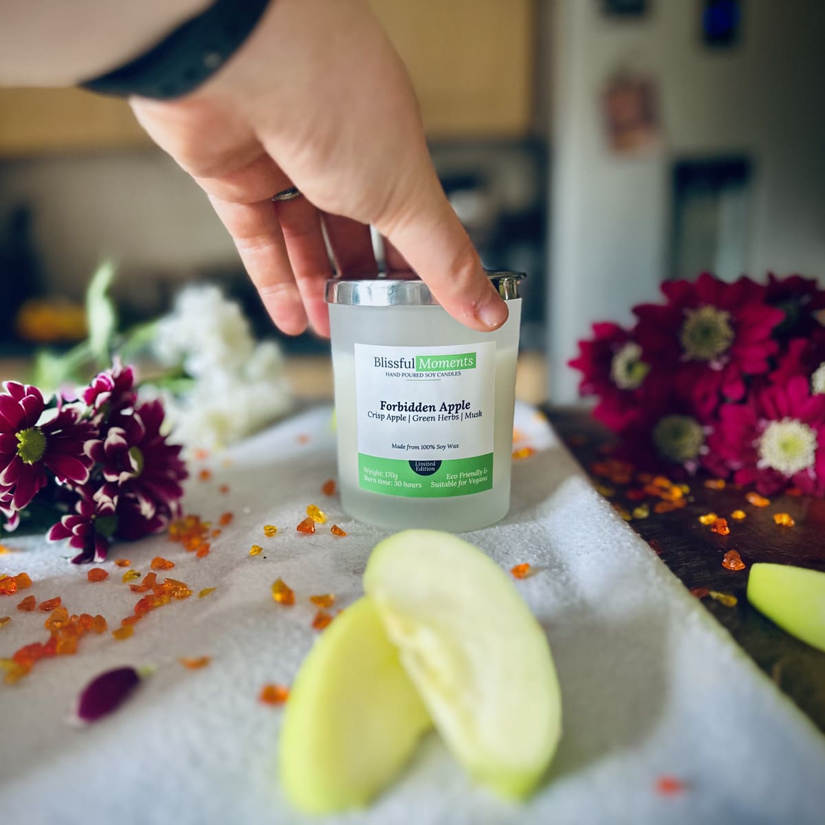 Forbidden Apple scented luxury frosted glass soy candle surrounded by fresh flowers, amber and green apples sitting on a cosy blanket