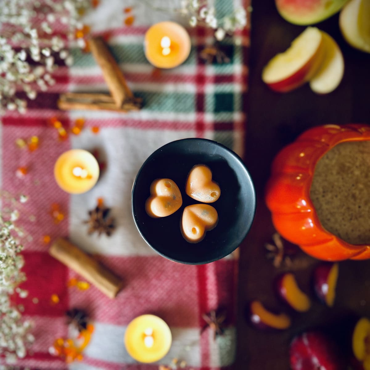 Three Orange Frosted Pumpkin scented heart shaped wax melt in a black wax melt burner dish on a festive table with candles, cinnamon and pumpkins.