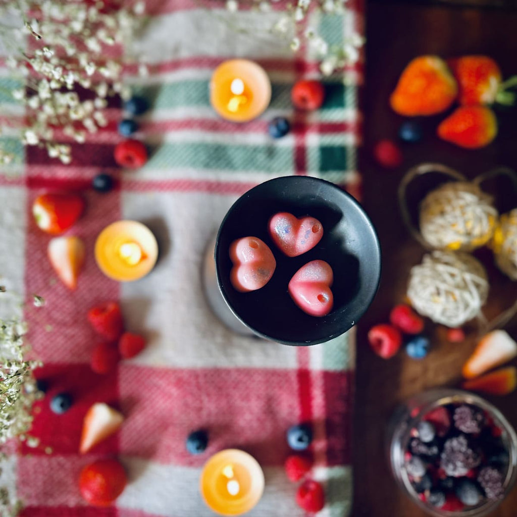 Pink coloured Frosty Berries scented Heart-shaped wax melts in a black wax melt burner dish on a checkered cloth surrounded by winter fruits and tealights.