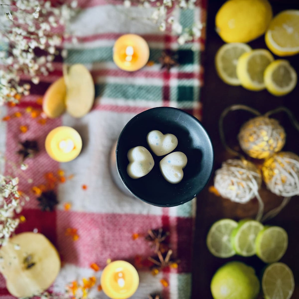3 white coloured Frosty Lake scented Heart-shaped wax melts in a black wax melt burner dish on a red festive plaid tablecloth with citrus fruits and Christmas lights