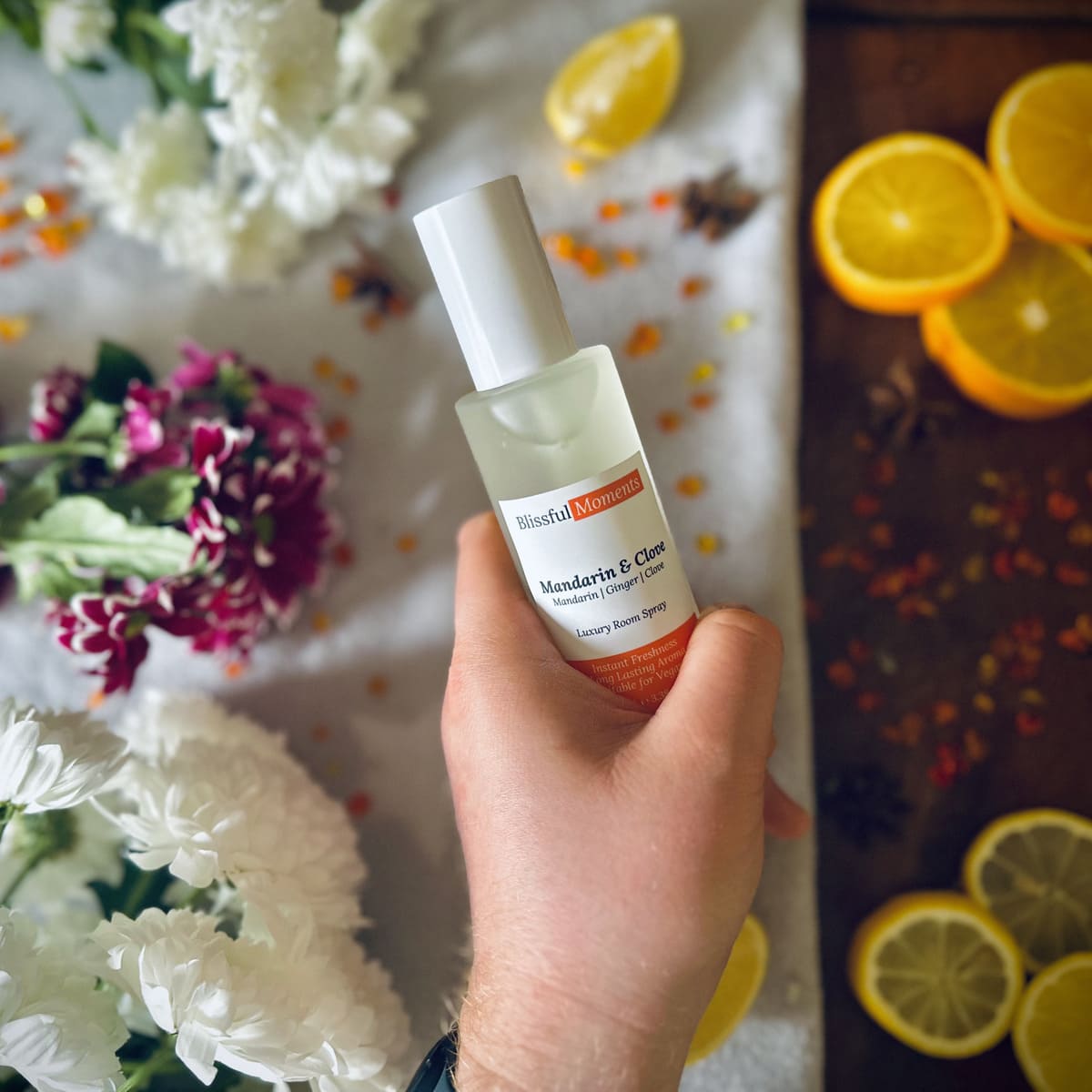Hand holding a luxury Mandarin & Clove scented frosted glass room spray surrounded by fresh flowers and citrus rooms sitting on a cosy blanket