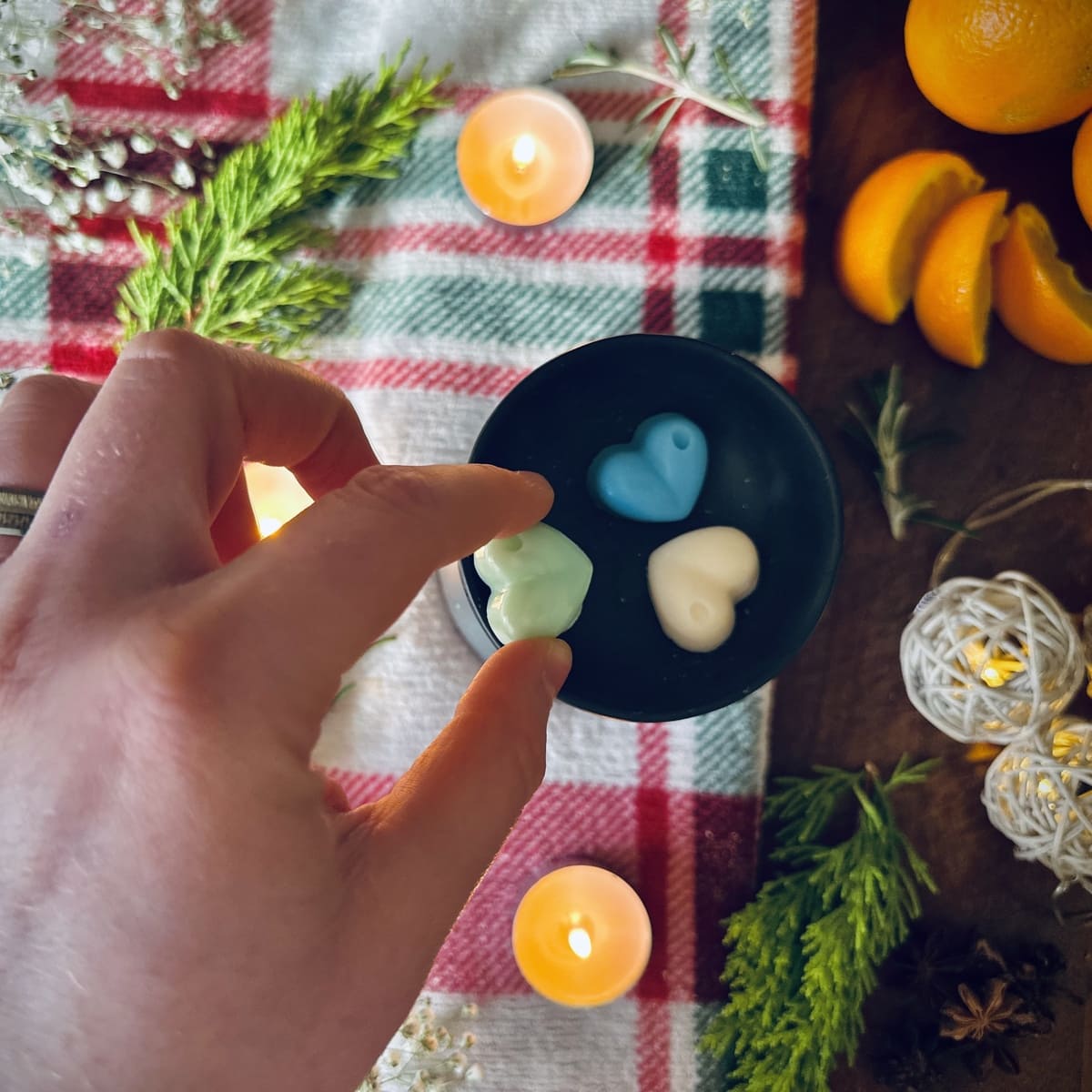 Hand holding a green coloured Northern lights heart shaped wax melt with a bue and white wax melt in a black wax melt burner on a festive background with candles and oranges.
