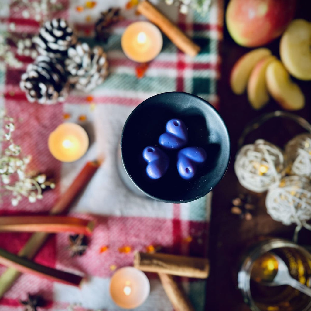 The purple coloured rhubarb and cinnamon scented heart shaped wax melts in a black wax melt burner on a festive tablecloth with tea lights and apples, rhubarb and cinnamon sticks.