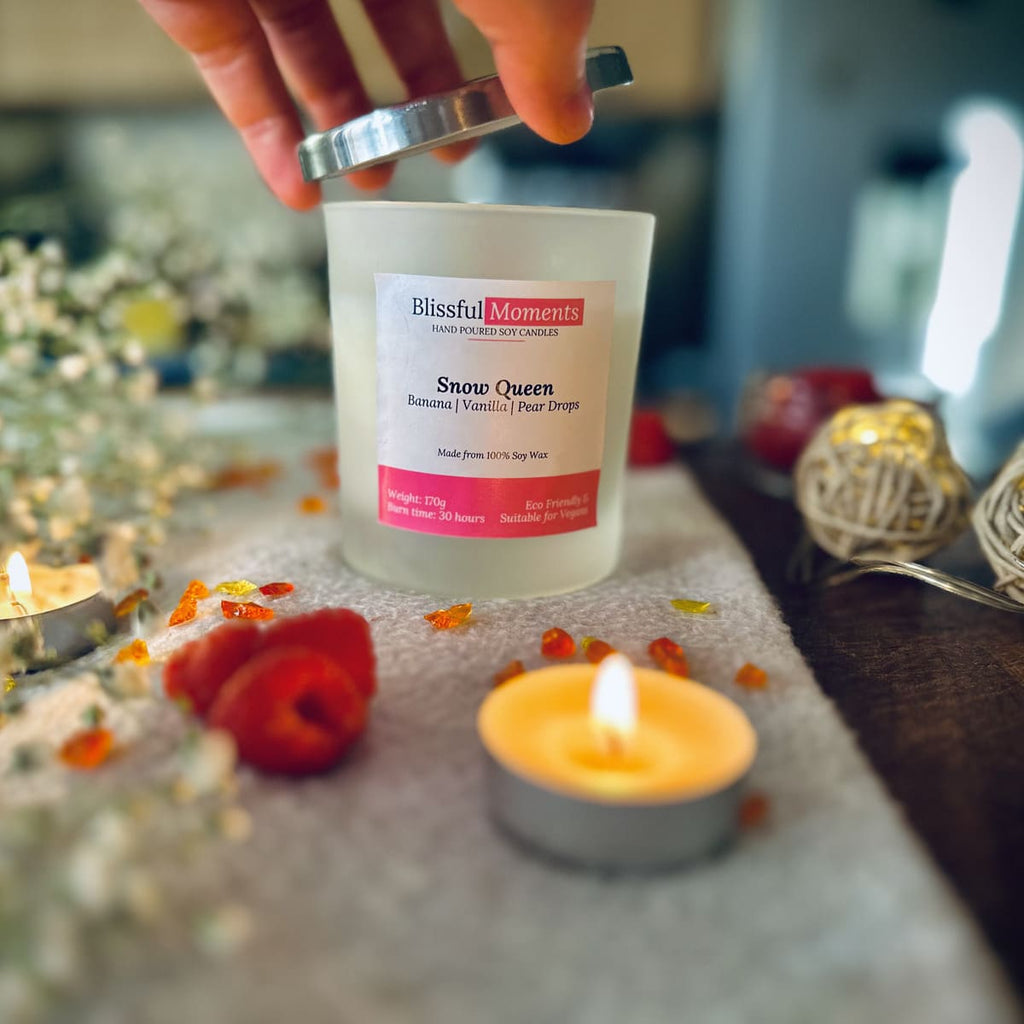 Hand taking the silver lid off the Snow Queen candle on a grey blanket surrounded by tea lights, raspberries and flowers