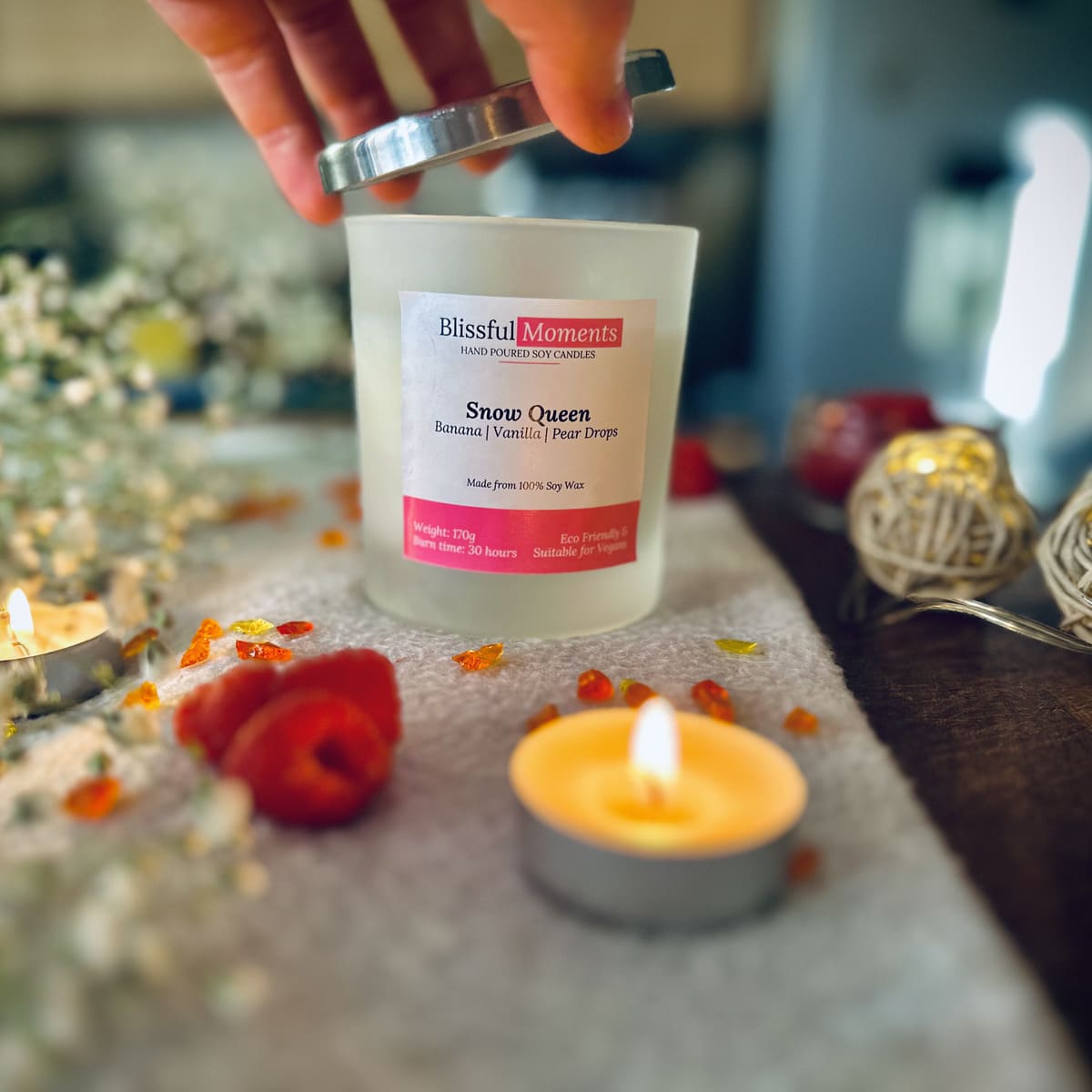 Hand taking the silver lid off the Snow Queen candle on a grey blanket surrounded by tea lights, raspberries and flowers
