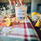 White Christmas scented soy candle with a hand removing the silver lid on a red checked tablecloth surrounded by christmas decorative items.