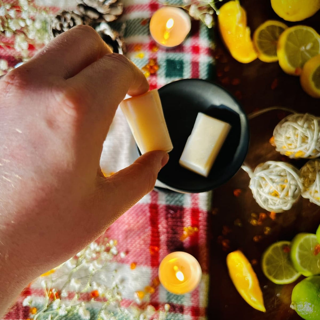 White Christmas snapbar wax melt segment being held by a hand and placing into a black tealight burner dish surrounded by lemons and candles on a Christmas checkered tablecloth.