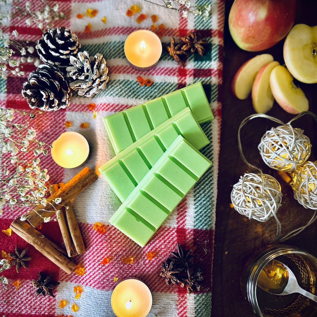 Green coloured Apple and Cinnamon scented snapbar wax melts on a Christmas checked plaid fabric with candles, apples, and festive decorative items.