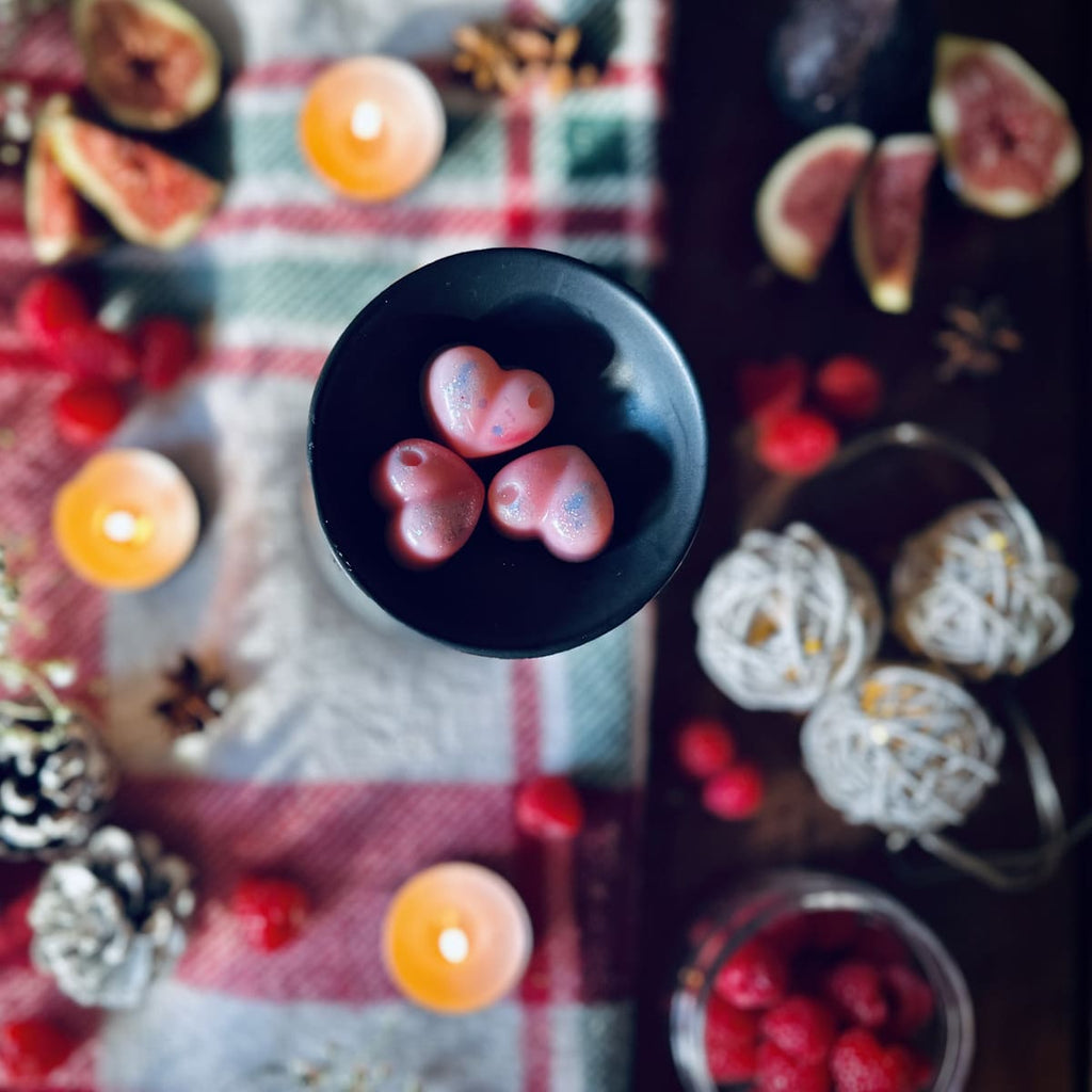 Three pink Cherry and Fig scented heart shaped wax melts in a black wax melt burner on a red festive checked tablecloth surrounded by figs, raspberries and christmas lights