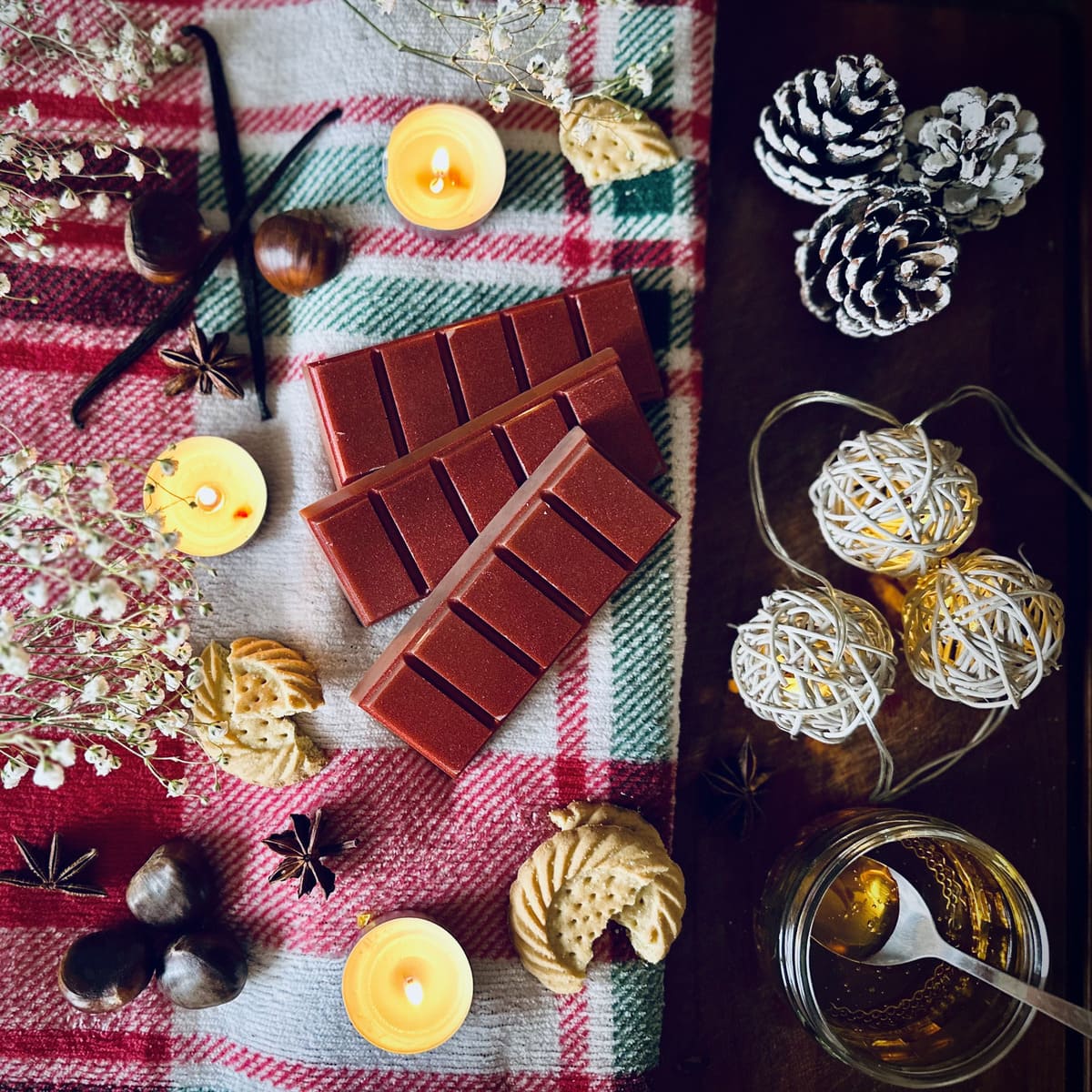 Red coloured Christmas Cookie scented snapbar wax melts on a checkered cloth surrounded by festive biscuits, chestnuts, caramel and pine cones.