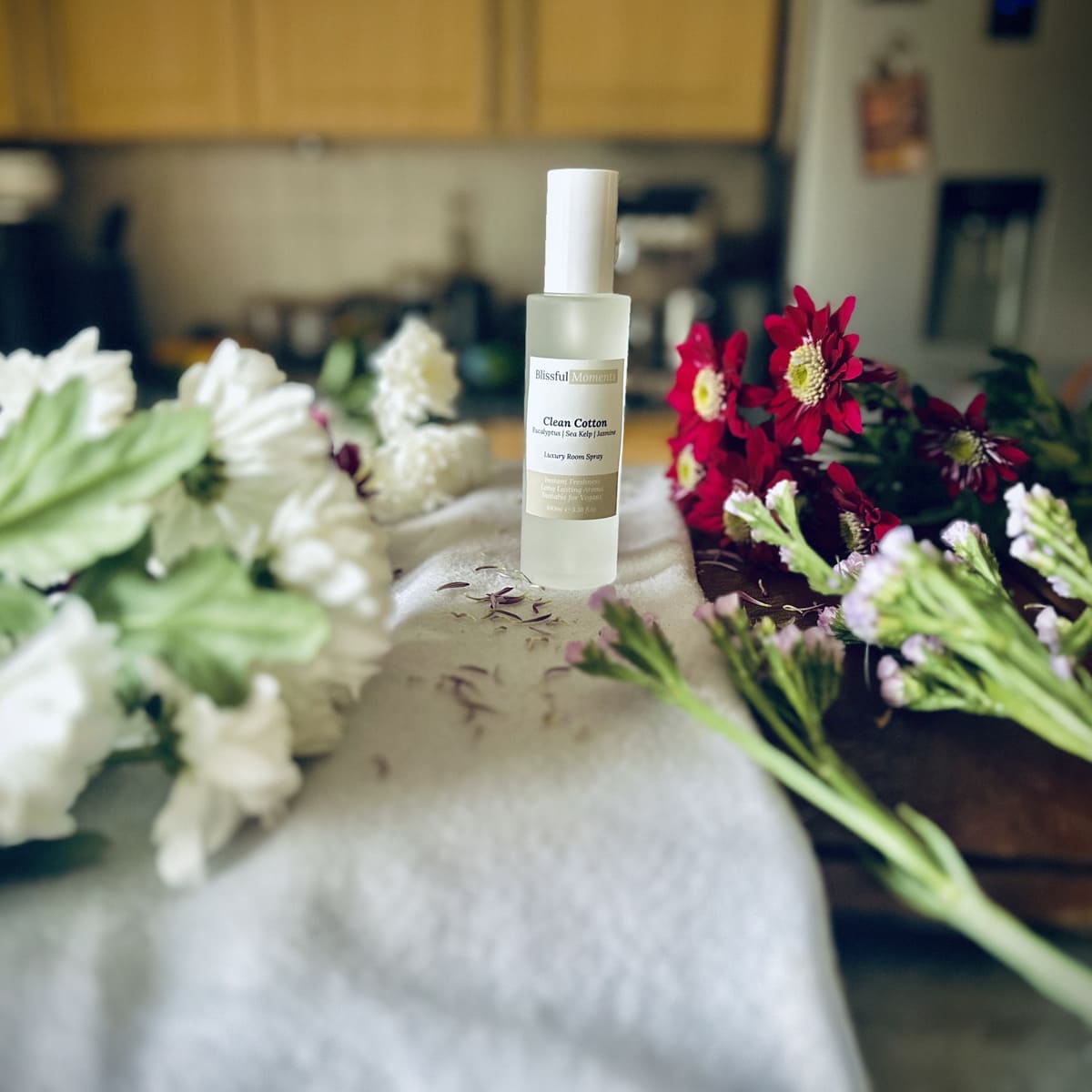 Luxury Clean Cotton scented room spray sitting on a soft cosy blanket surrounded by fresh flowers.