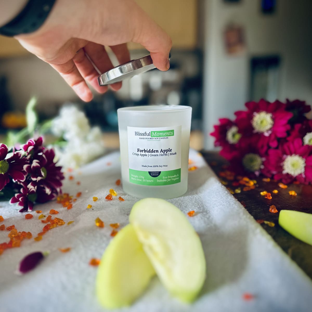 Forbidden Apple scented luxury frosted glass soy candle surrounded by fresh flowers, amber and green apples sitting on a cosy blanket