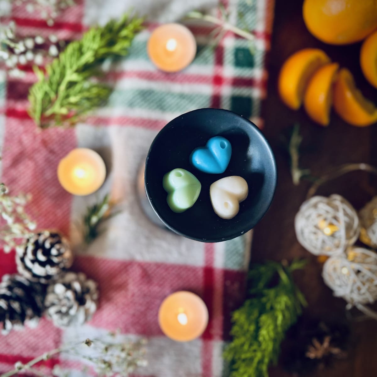 blue, green and white coloured northern lights scented heart shaped wax melts in a black wax melt burner on a red festive checked blanket surrounded by oranges, pinecones and rosemary.