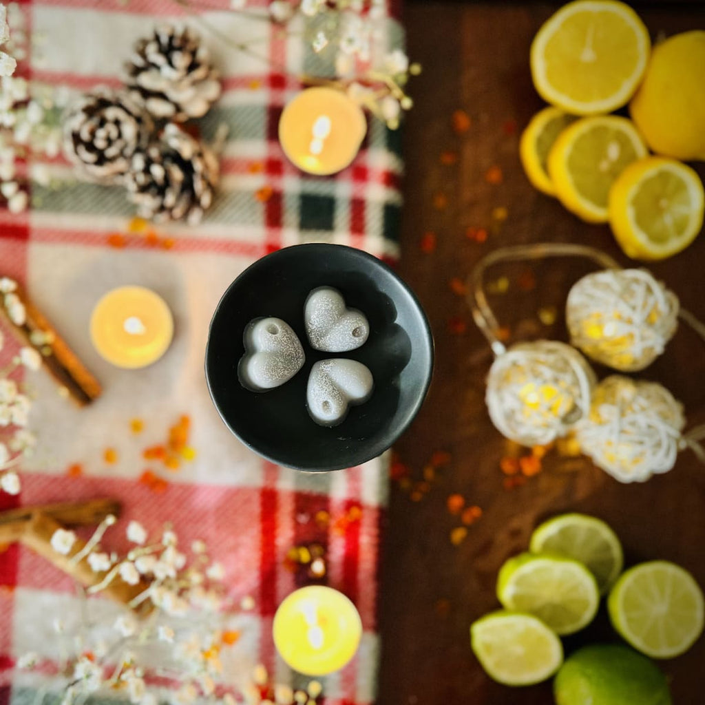 Grey Starry Night heart shaped wax melts in a black tealight burner with a Christmas Decorative setup with candles, lemons, limes, and pinecones on a checkered tablecloth.