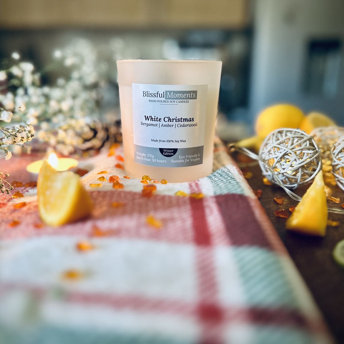 White Christmas scented soy candle on a checked tablecloth surrounded by amber, oranges and christmas lights