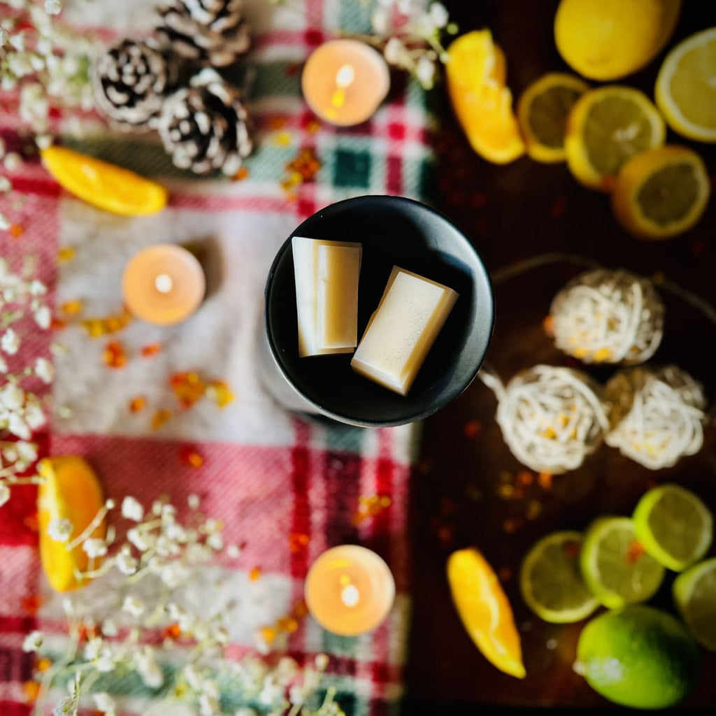 Two White Christmas snapbar wax melt segment sitting in a black tealight burner dish surrounded in a decorative Christmas set up with lemons, limes, and candles on a festive checkered tablecloth