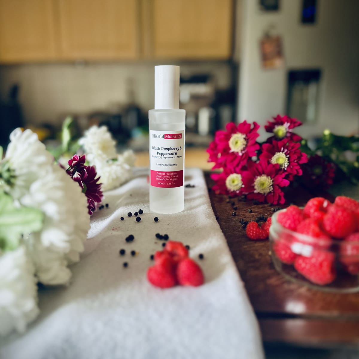 Black Raspberry & Peppercorn scented luxury frosted glass room spray surrounded by fresh flowers, peppercorns and raspberries sitting on a cosy blanket