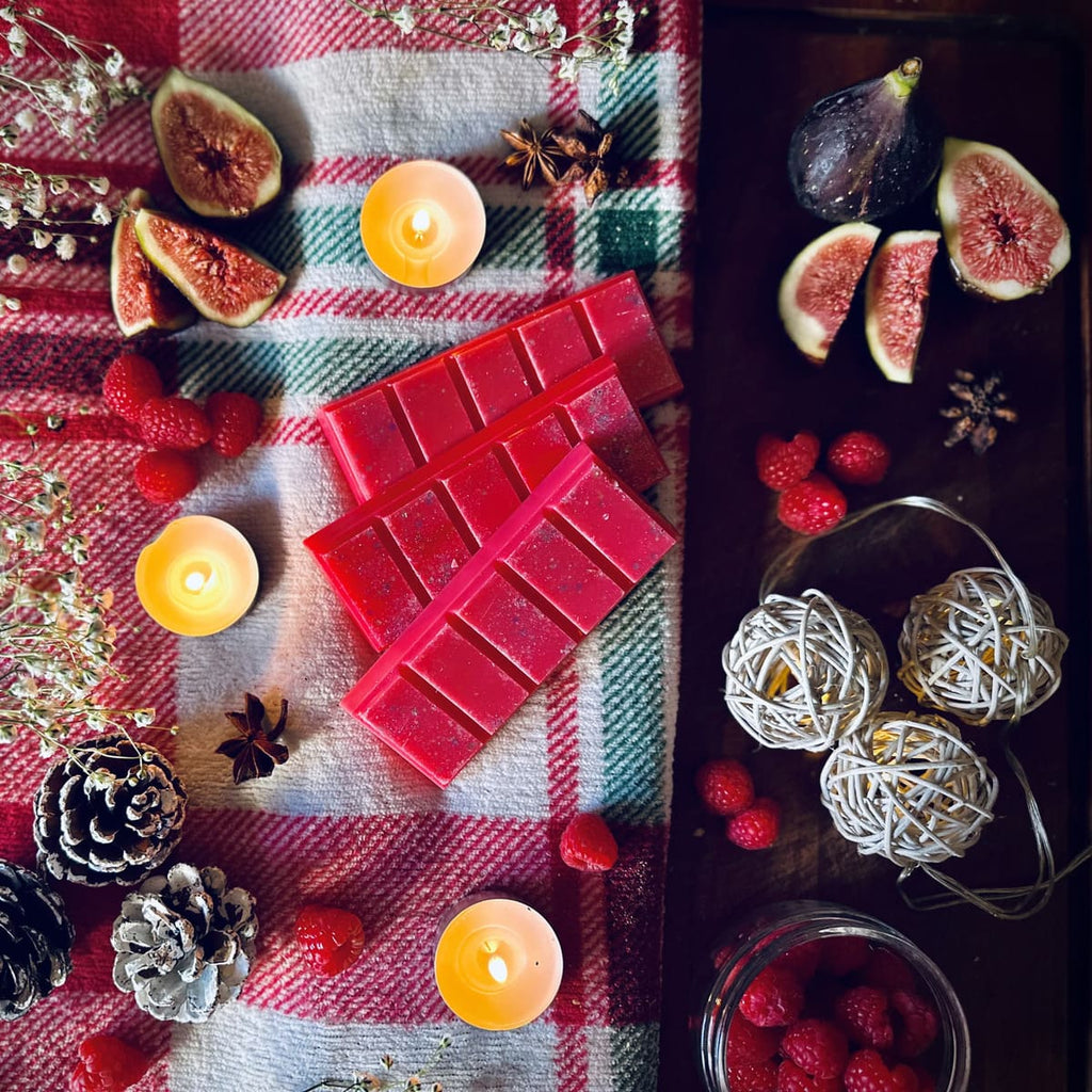 Red coloured Cherry and Fig scented snapbar wax melts on a festive checked tablecloth surrounded by figs, raspberries, pinecones and Christmas. lights.