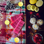 Red Christmas Wish scented snapbar wax melts on a checkered cloth with candles and berries, and lemons and berries on a dark surface.