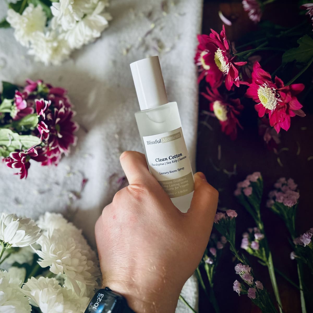 Hand holding a luxury Clean Cotton scented room spray surrounded by fresh flowers.