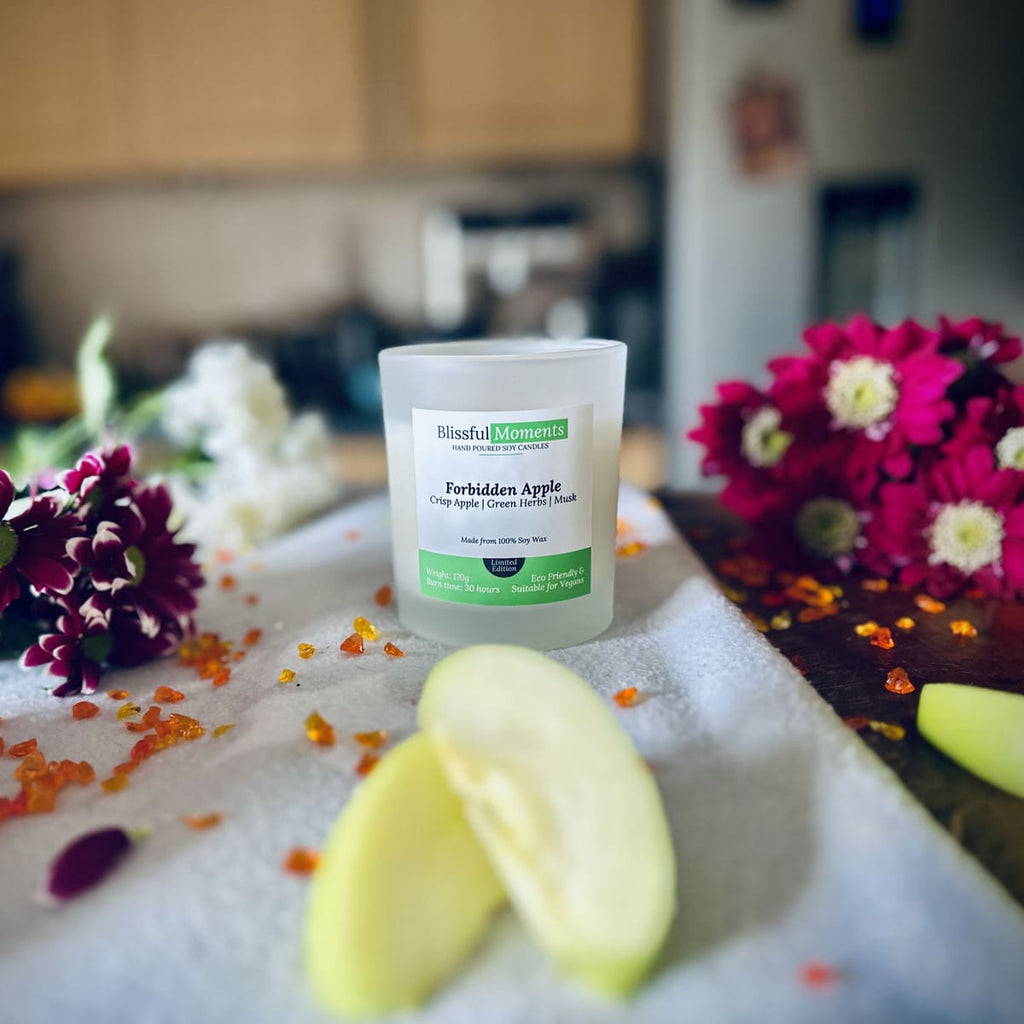 Forbidden Apple scented luxury frosted glass soy candle surrounded by fresh flowers, amber and green apples sitting on a cosy blanket