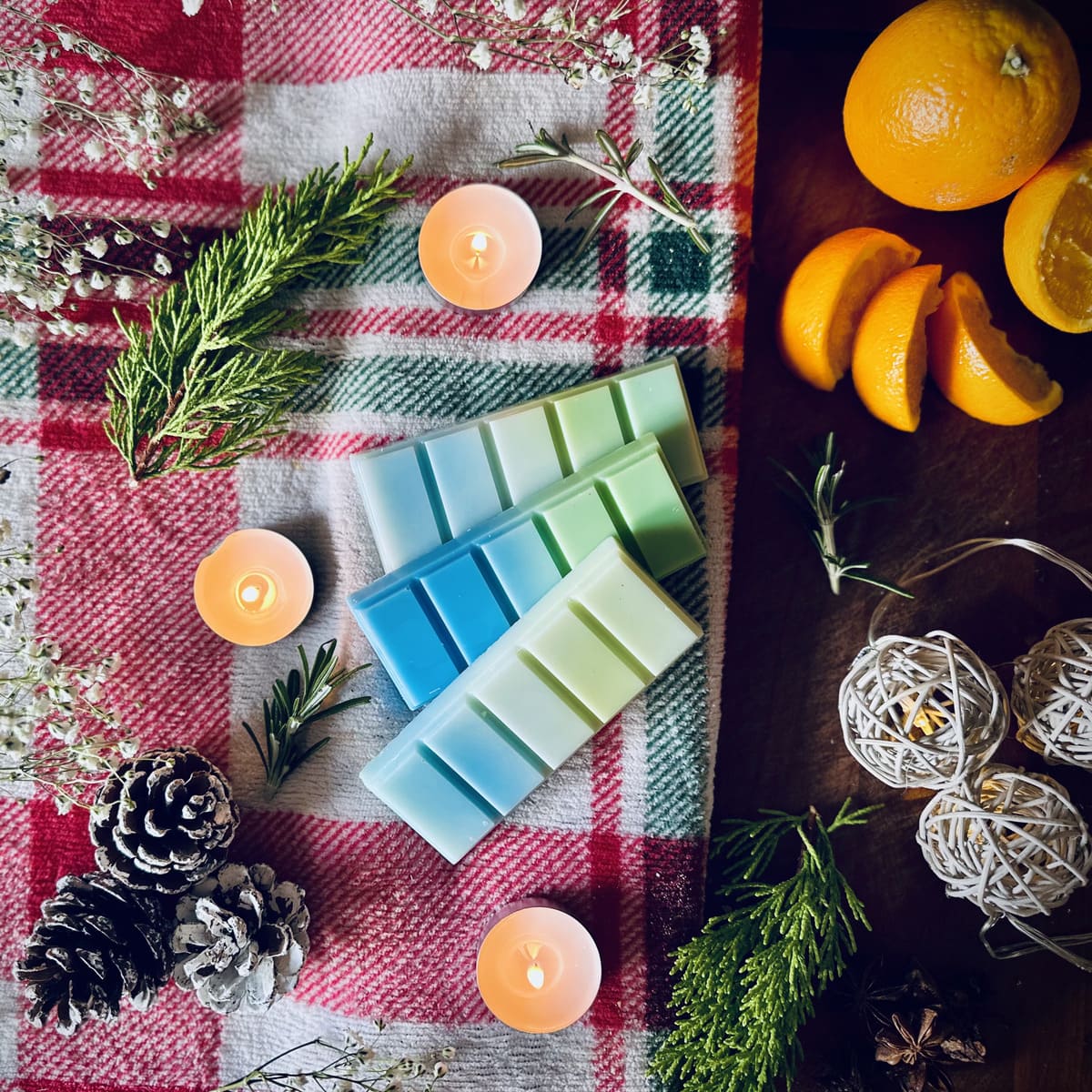 Northern lights scented snapbar wax melts on a Christmas checkered cloth with tea lights, pinecones, oranges and rosemary