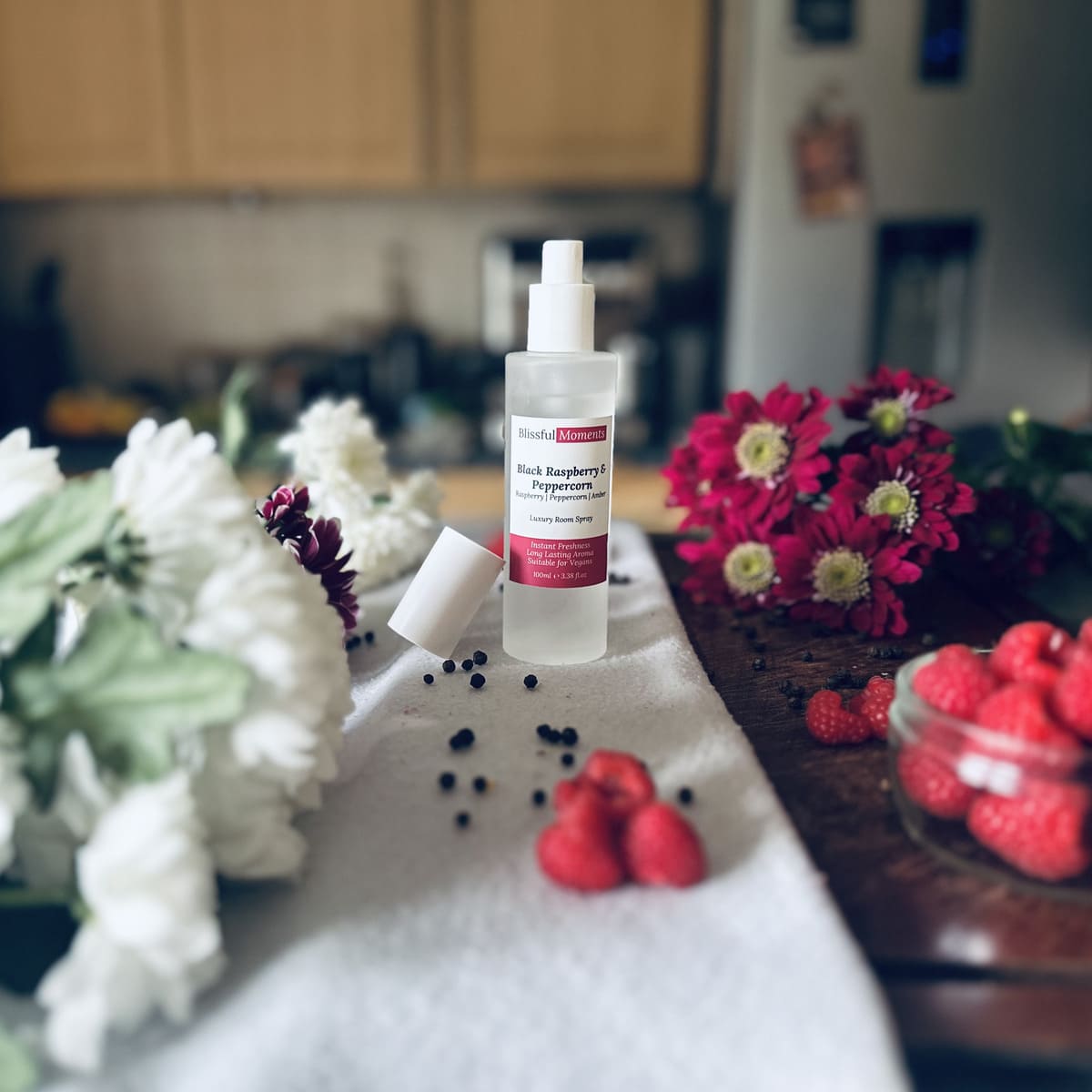 Black Raspberry & Peppercorn scented luxury frosted glass room spray surrounded by fresh flowers, peppercorns and raspberries sitting on a cosy blanket