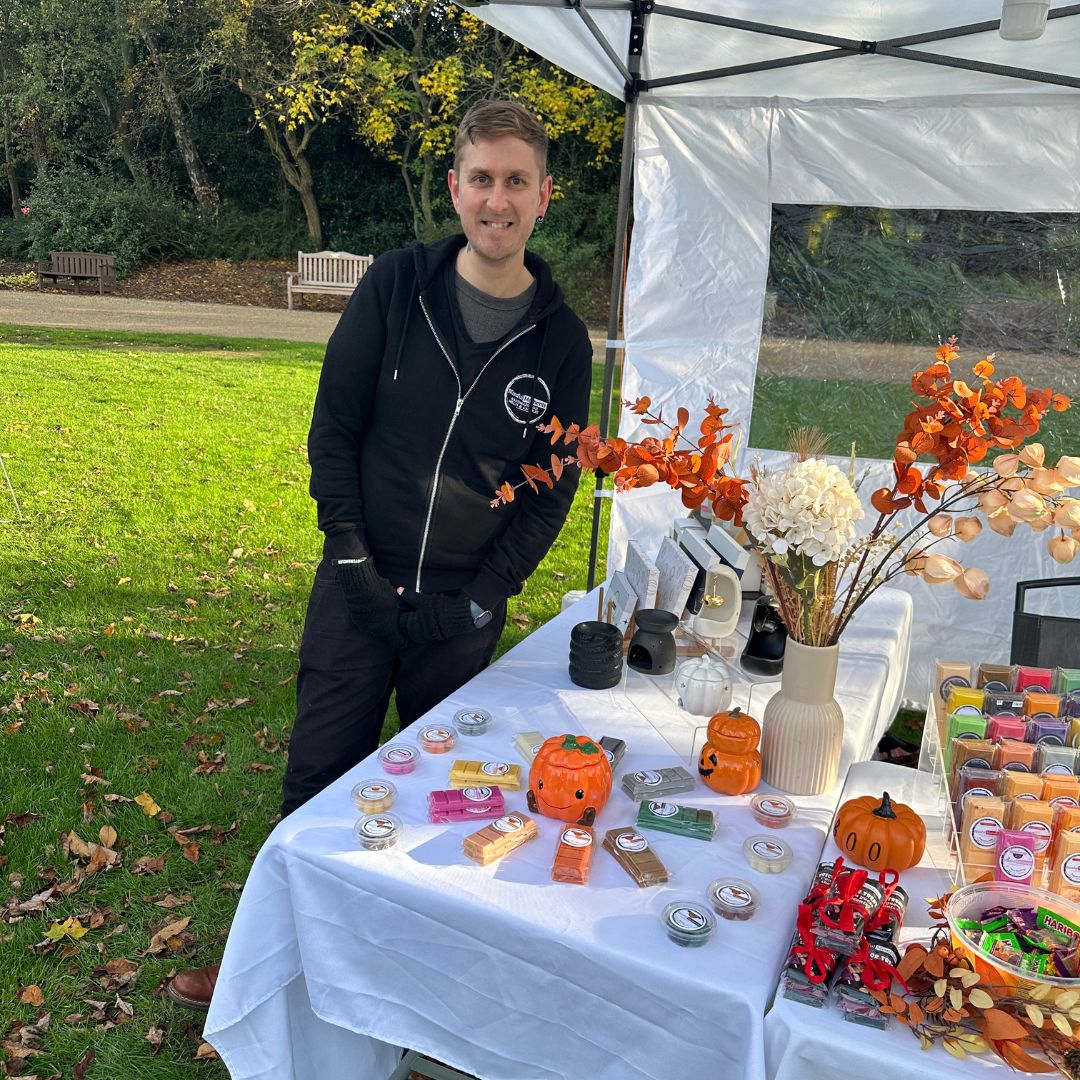 A man in a black zip-up hoodie stands behind an outdoor craft table set up under a white canopy in a park. The table is covered with various handmade items, including small orange jack-o'-lantern candles, colorful wax melts or soaps, and decorative autumn branches.