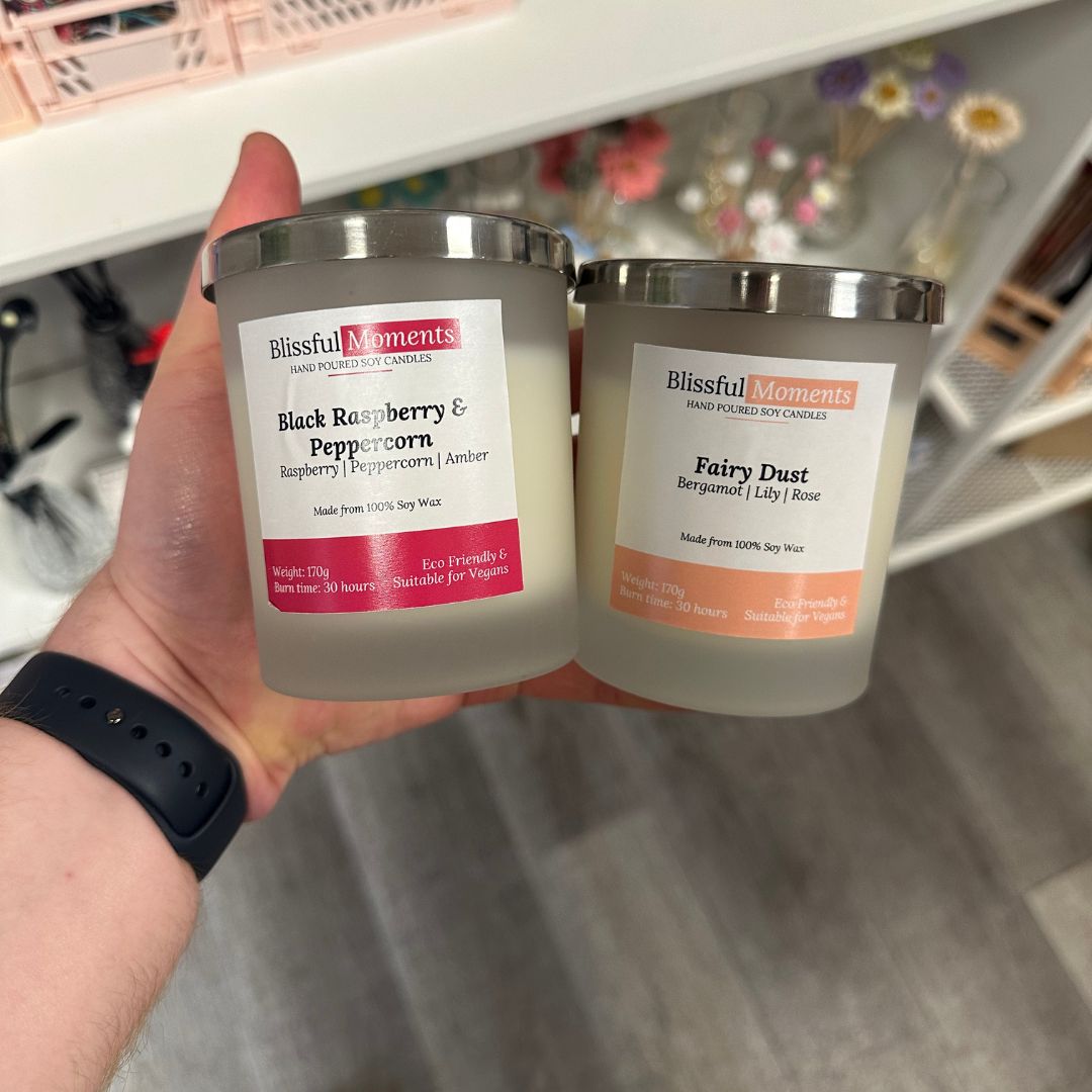 A hand holds two Blissful Moments scented soy candles in frosted glass jars with silver lids. The left candle is labeled 'Black Raspberry & Peppercorn' with a pink label, and the right candle is labeled 'Fairy Dust' with an orange label.