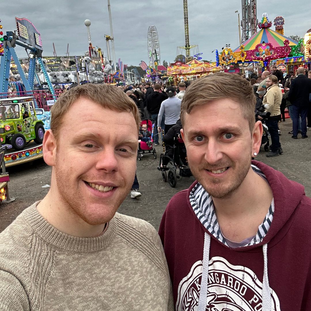 Tom and Hasip at Hull Fair - Two men smiling for a selfie at a busy outdoor fairground with carnival rides and attractions in the background.