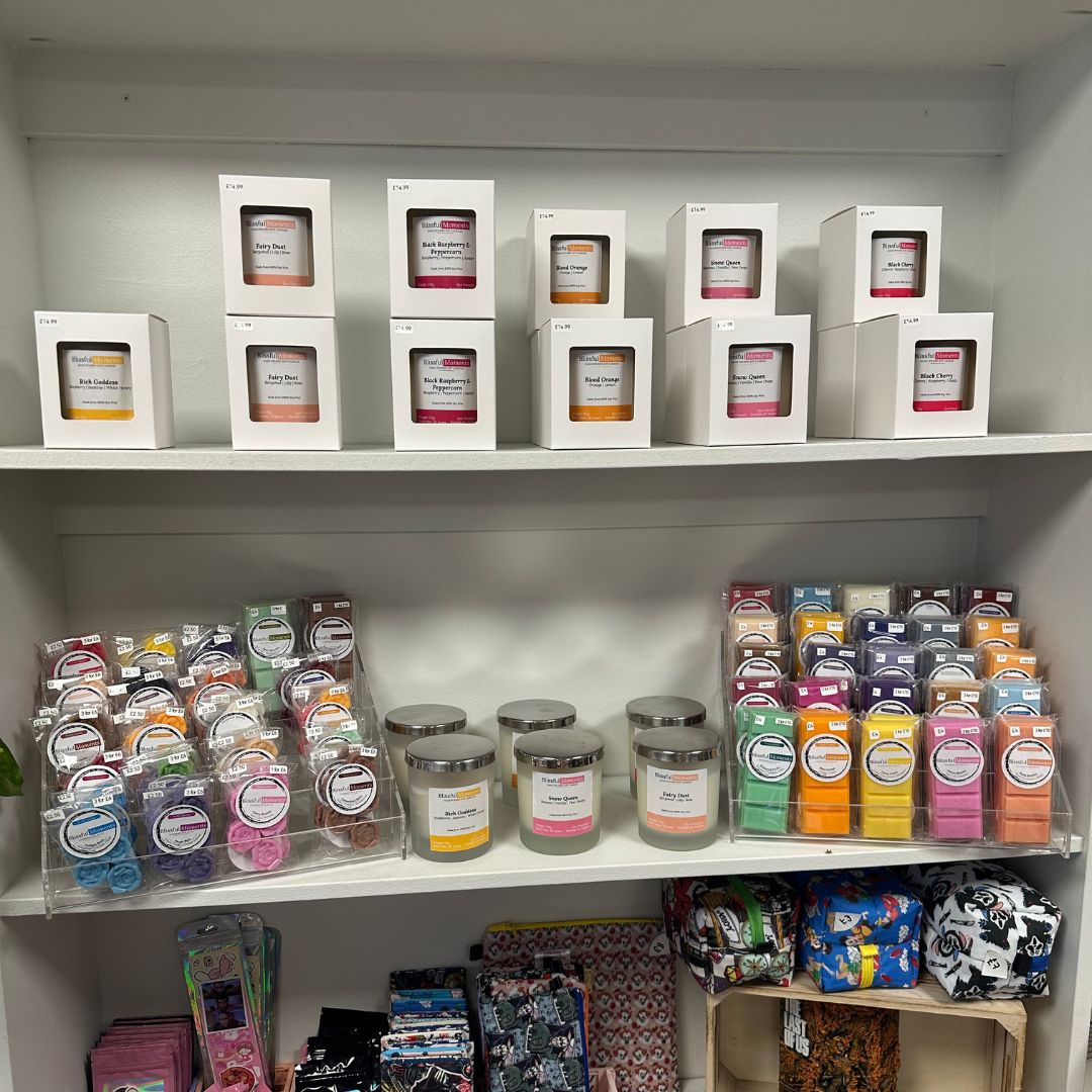 A retail display shelf filled with various scented candles in different packaging styles, including white boxes with clear windows, glass jars, and colorful individually wrapped wax melts.