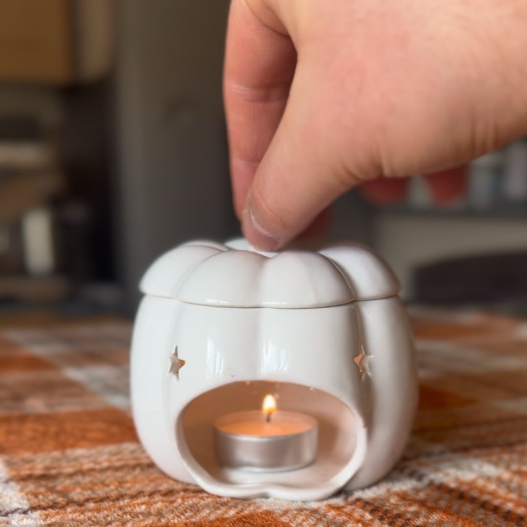 Why choose Blissful Moments - A white ceramic pumpkin-shaped tea light holder with star cutouts, containing a lit tealight candle, with a hand lifting the lid in a cozy indoor setting.